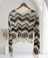 Blusa de manga larga tejida a crochet, con patrón de zigzag en tonos marrón oscuro y beige. Presenta motivos circulares en el ruedo y terminaciones con borlas colgantes.
