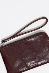 Monedero de piel color marrón rojizo con acabado brillante, estructura rígida y cierre de cremallera con tirador de tejido. Presenta logo metálico de Bimba y Lola en el frente y bolsillo interior.
