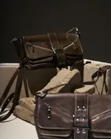 Cartera bandolera marrón con correa ajustable, cierre con hebilla y detalles de cremalleras decorativas.