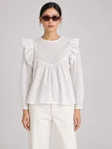 Blusa blanca de algodón con detalles de bordado broderie, cuello redondo y mangas largas abullonadas. Presenta volados decorativos en los hombros y diseño holgado.