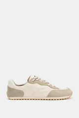 Zapatillas deportivas estilo barefoot con puntera amplia y suela plana flexible. Presentan un diseño combinado en tonos beige y crema, con paneles de gamuza y tela, cierre mediante cordones bicolor y suela con efecto engomado.