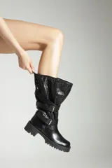 Bota de caña alta color negro, confeccionada en una combinación de cuero liso en la base y material tipo mesh perforado en la caña. Presenta múltiples correas horizontales con hebillas metálicas decorativas y una suela track de goma con taco bajo.