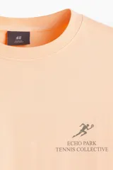 Remera color durazno de corte holgado con cuello redondo y estampado pequeño en el pecho con un tenista y texto.