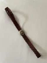 Cinto trenzado de cuero color chocolate con hebilla circular metálica grabada y piedra central.