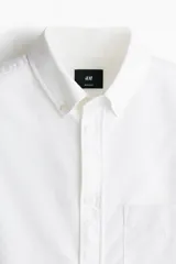 Camisa Oxford de algodón blanca, de corte regular fit, con cuello americano, bolsillo frontal aplicado y mangas largas con puños abotonados.