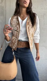 Chaleco beige con bordados blancos en estilo ornamental, sin mangas y con cierre frontal abierto.