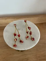 Aretes colgantes largos, con estructura de metal dorado (bronce) que simula ramas o tallos, de los cuales cuelgan irregularmente pequeñas piedras o apliques de color rojo intenso con detalles de pan de oro o láminas doradas incrustadas.
