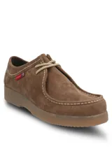 Zapato tipo wallabee de Hush Puppies, confeccionado en cuero gamuzado color marrón. Presenta costuras a la vista en tono claro, cordones al tono y suela de goma texturizada. Incluye etiqueta lateral con la marca y bandera británica.