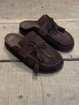 Zueco tipo mocasín de gamuza color marrón chocolate, con detalle de costuras visibles en el borde y cordones decorativos con borlas en el empeine.