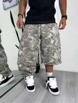 Pantalón ancho tipo baggy de tela estampada con patrón de camuflaje abstracto en tonos grises y beige. El largo es a media pierna, estilo bermuda ancha, con puños doblados.