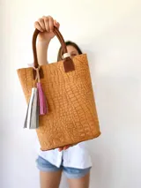 Bolso tipo tote de cuero color marrón con textura tipo cocodrilo, doble asa corta de cuero marrón y detalle de borlas decorativas.