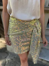 Falda corta de lentejuelas con diseño cruzado y detalle de lazo lateral. Presenta un acabado brillante con destellos iridiscentes.