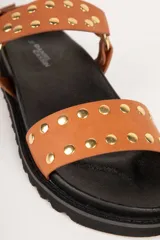 Sandalia de mujer color marrón, estilo pescador o chunky, con plataforma negra dentada. Presenta dos tiras anchas adornadas con tachas doradas y una tira de ajuste en el talón con hebilla metálica dorada.