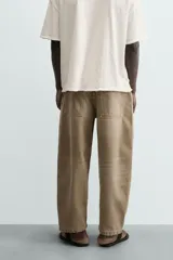 Jeans de hombre, tiro medio, color marrón claro o caqui, con corte relaxed fit y perneras anchas. Presenta cinco bolsillos y costuras laterales giradas hacia el frente.