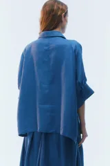 Camisa oversize blanca de lino, con corte cuadrado, mangas raglán cortas con puños anchos, aberturas laterales y cierre frontal de botones.