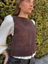 Chaleco de cuero marrón chocolate, sin mangas, con escote redondo y detalles de costuras decorativas en forma de espiga en el frente. Cuenta con cordones laterales para ajustar.