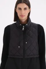 Chaleco largo negro de diseño combinado, con parte superior en matelassé y parte inferior de tela lisa. Presenta cuello alto con cordones ajustables y cierre frontal completo.