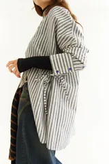 Camisa oversize de corte holgado con estampado de rayas verticales. Presenta cuello camisero, cierre frontal con botones a contraste y cordones integrados en la cintura para ajustar la silueta. Posee mangas largas con puños rectos.