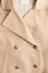 Gabardina corta color beige, de estilo cruzado con doble botonadura, solapas de muesca, bolsillos ribeteados y mangas raglán con charreteras en los hombros.