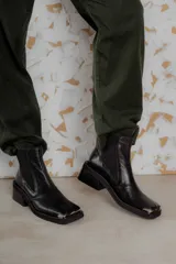 Botas tipo chelsea de cuero color marrón oscuro con acabado brillante. Presentan punta cuadrada con detalle de puntera metálica, elásticos laterales y taco cuadrado bajo.