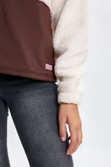 Buzo de cuello alto con medio cierre frontal, confeccionado en una combinación de tejido tipo sherpa en la parte superior y nylon en la parte inferior. Presenta un diseño bicolor en tonos crudo y marrón, con detalles de cierre en color rosado.