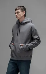 Hoodie oversize color gris oscuro, confeccionado en frisa premium con textura gamuzada. Presenta capucha ajustable con cordones personalizados, bolsillo frontal tipo canguro y puños con abertura para el pulgar.