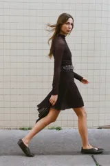 Vestido de tejido de punto marrón oscuro, con cuello alto, manga larga y bajo asimétrico con volado. Se complementa con un cinturón ancho negro con tachas circulares y hebilla metálica.