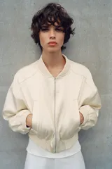 Chaqueta bomber corta de tejido satinado color beige, con cuello redondo, cierre frontal con cremallera, mangas largas de hombro caído y bolsillos de vivo en el delantero. Presenta acabados en rib elástico en cuello, puños y ruedo.