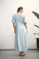 Vestido largo color beige claro, con escote en V, manga corta acampanada y cierre frontal de botones. Presenta un lazo ajustable en la cintura y un tejido con textura sutil.