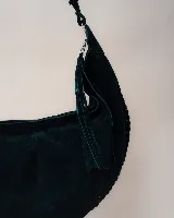 Bolso tipo hobo de gran tamaño, confeccionado en gamuza color marrón oscuro. Presenta un diseño artesanal con costuras visibles y detalles de tientos en el cuerpo, asa integrada con pespuntes a mano y cierre superior.