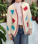 Cardigan de lana merino tejido a mano con técnica de dos agujas, de color marfil con parches geométricos de tela en tonos celestes y lila aplicados con costuras visibles. Presenta cierre frontal con botones de madera y detalles de ribetes multicolores en los bordes.