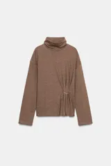 Top de cuello alto y manga larga, confeccionado en tejido de algodón gris melange. Presenta un diseño con frunces laterales ajustados mediante un aplique metálico decorativo.