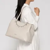 Cartera tipo tote de cuero sintético color gris, con doble asa de hombro y cadena colgante plateada con dijes de letras que forman la marca. Presenta el logo de Guess en la parte frontal y herrajes plateados.