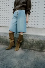 Bota de caña alta color verde oliva, confeccionada en serraje, con pespuntes decorativos y tacón tipo kitten. Acabado en punta.