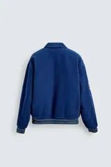 Cazadora estilo varsity confeccionada en pana de algodón azul, con interior ligeramente acolchado. Presenta cuello solapa, cierre frontal con botones a presión, bolsillos laterales de vivo y acabados en rib a rayas en puños y cintura. Incluye múltiples parches bordados decorativos en el frente.