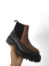Botín tipo chelsea de cuero con diseño bicolor, combinando paneles en color marrón texturizado y gris oscuro liso. Cuenta con elásticos laterales negros y suela track robusta de goma.