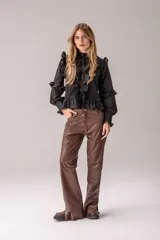 Camisa negra de corte entallado con volados decorativos en el cuello, la botonadura frontal, los hombros y las mangas. Presenta mangas largas con puños ajustados y un ruedo con volado inferior.