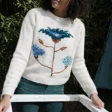 Sweater de punto de lana merino color blanco crudo, con cuello redondo, mangas largas y un diseño tejido y bordado a mano de una gran flor en la parte frontal.