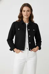 Chaqueta de jean negra con cuello camisero, cierre frontal con botones metálicos y mangas con sutiles frunces en los hombros. Presenta un corte entallado al cuerpo y bolsillos frontales con solapa.