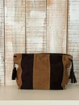 Bolso tipo sobre y bandolera de gamuza en tonos marrones y tostados, con diseño de franjas verticales y borlas colgantes en los laterales.