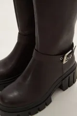 Bota de caña alta color negro, confeccionada en eco cuero, con suela track de goma y detalle de hebilla metálica en el tobillo y en la parte superior de la caña.