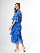 Vestido midi de seda italiana color azul, con escote en V, mangas murciélago amplias, falda de tres volados y cintura elastizada con lazo ajustable al tono y hebilla circular dorada.