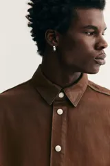 Camisa de corte oversized en denim de algodón rígido revestido, color marrón oscuro. Presenta cuello inglés, tapeta clásica con botones de metal y canesú en la espalda. Tiene hombros caídos, mangas largas con puños abotonados y bajo redondeado.