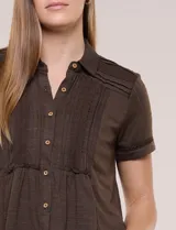 Camisa de manga corta color marrón oscuro, con cuello camisero, alforzas verticales, detalles de puntilla calada y frunces bajo el busto. Cuenta con cierre frontal de botones.