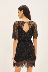 Vestido corto negro con mangas cortas de encaje, bordado a mano con pequeñas cuentas o lentejuelas. Presenta un escote pronunciado en forma de U con un corte de gota central y un cinturón fino bordado a juego. El ruedo es de encaje con terminación festoneada.
