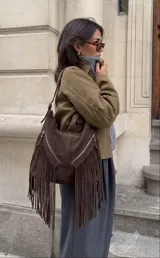 Bolso tipo hobo de cuero gamuzado color marrón oscuro, con diseño de flecos largos en el frente y dos bolsillos diagonales con cierre metálico. Cuenta con manija regulable y cierre principal metálico.