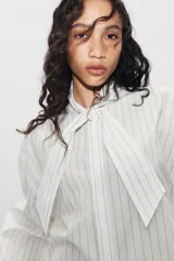 Camisa de popelín de algodón a rayas verticales, con cuello subido y aplicación de tejido tipo pañuelo anudado al frente. Presenta manga larga con puños anchos y cierre frontal con botones ocultos.