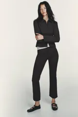 Pantalón entallado de tiro alto color negro, con cintura con trabillas y bolsillos de vivo con cremallera en el delantero. El bajo tiene un acabado en línea evasé. Cierre frontal con cremallera, botón interior y gancho metálico.