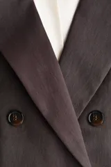 Blazer cruzado marrón oscuro con solapas de pico, hombreras y cierre frontal de doble botonadura. Cuenta con bolsillos con solapa en la parte delantera, puños con botones y forro interior.