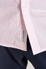 Camisa de manga larga confeccionada en tejido poplin con diseño de rayas verticales finas en color rosado y blanco. Presenta cuello abotonado (button-down), cierre frontal con botones y un bolsillo en el pecho.